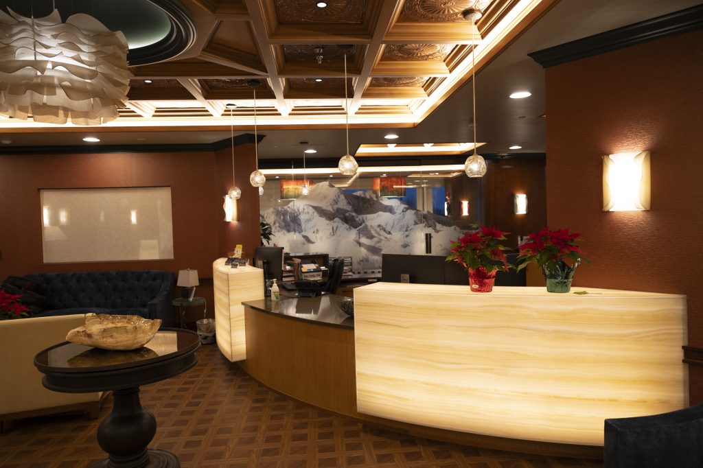 A photo of Northstarr CTS' reception area. A front desk is visible on the right, with a table and window on the left side of the frame. The room features warm colors and textures (wood desk, burgundy walls), and an elegant paper chandelier hangs from the ceiling.